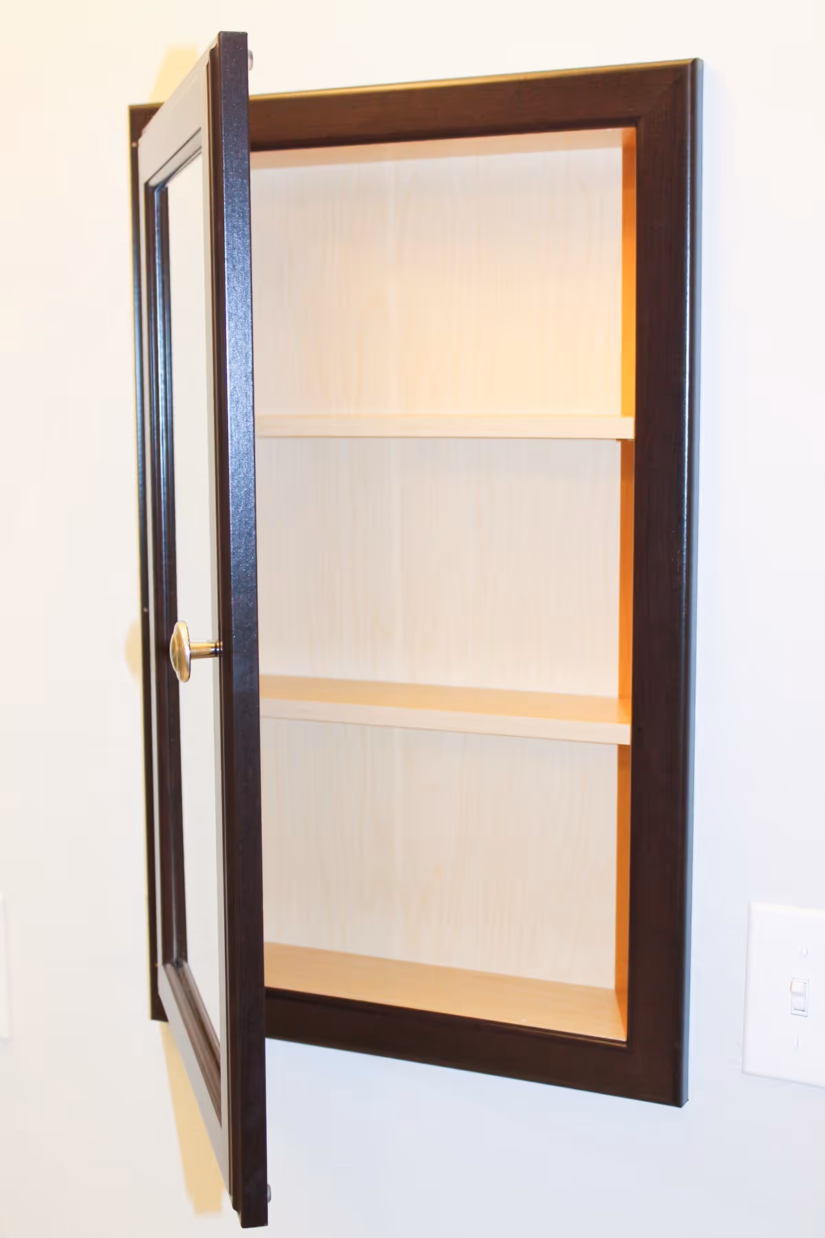 A wall-mounted wooden medicine cabinet with a dark brown frame and a glass door that is open, revealing two empty shelves inside. The cabinet is installed on a light-colored wall next to a light switch.