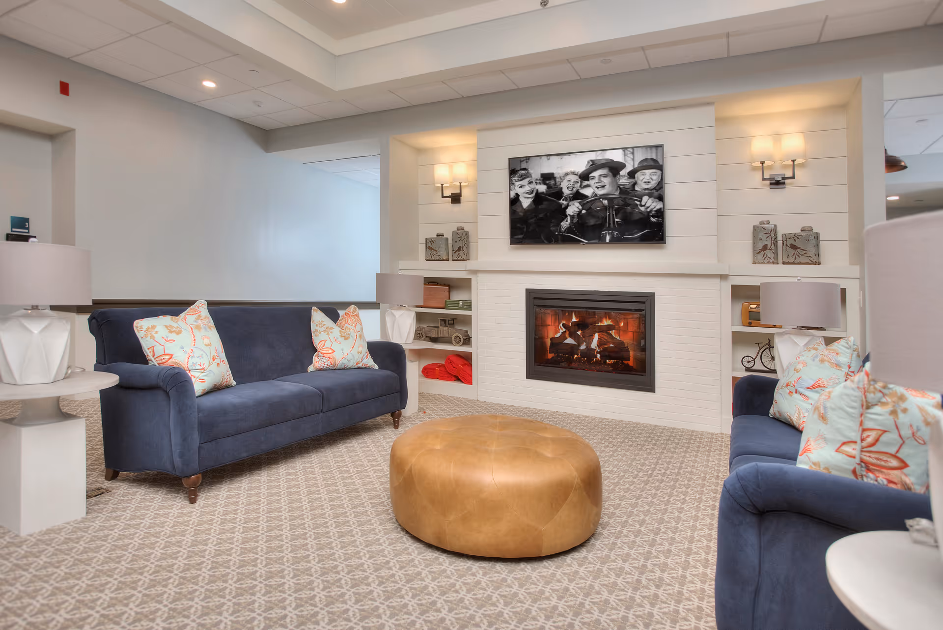 Communal living room with navy sofas, patterned pillows, a round leather ottoman, and a fireplace with a TV above.