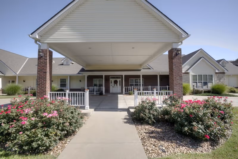 Front exterior view of a senior living facility with a covered entrance supported by brick pillars, a concrete walkway leading to double doors, and landscaped flower beds with pink flowers on either side.