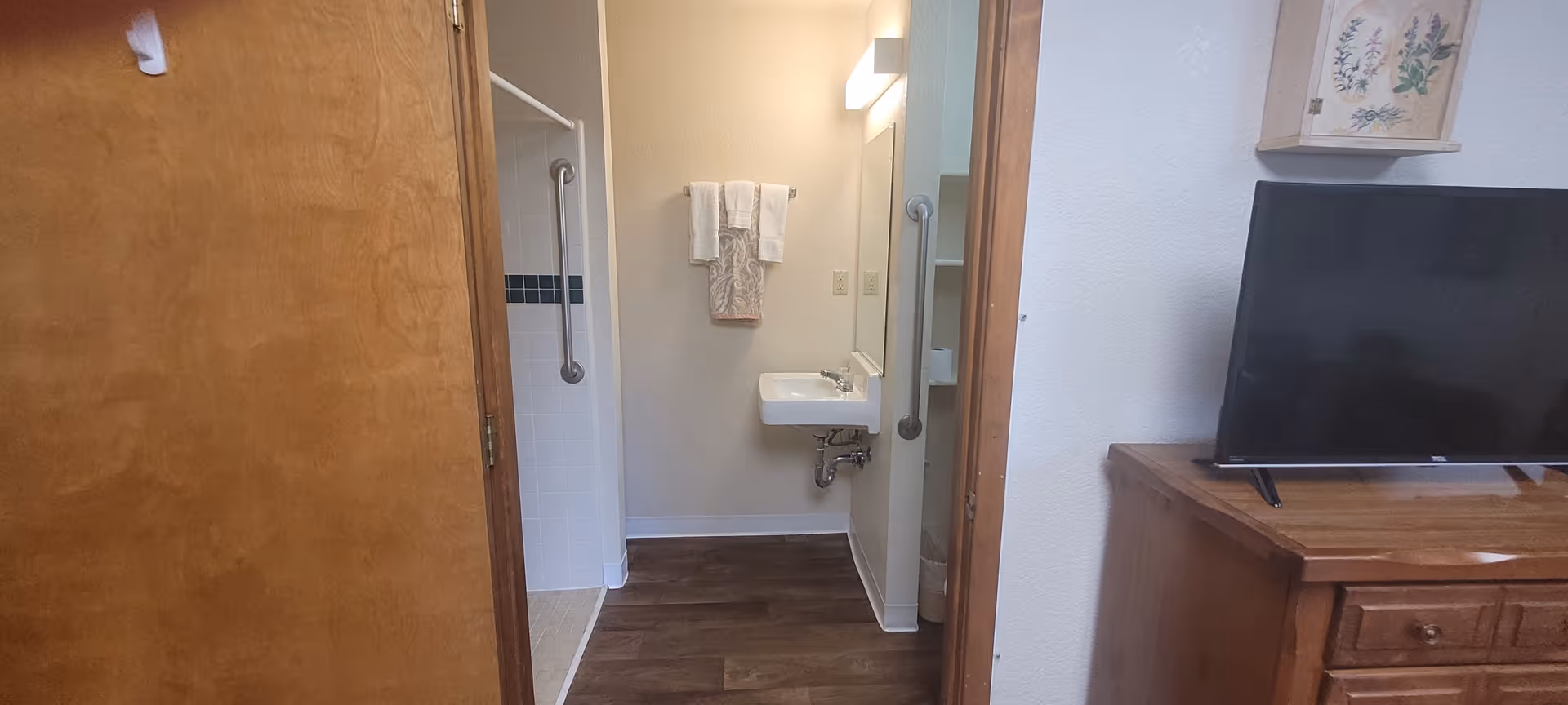 View through a doorway into a bathroom with a small sink, a mirror, and a towel rack holding three towels. To the left is a shower area with a grab bar and white tiles with a blue accent strip. To the right, part of a wooden dresser with a flat-screen TV on top is visible, along with a wall decoration above it.