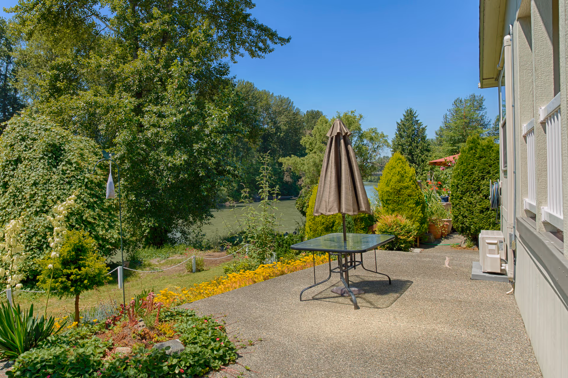Patio with a glass table and closed umbrella overlooking a landscaped yard, trees, and a lake.
