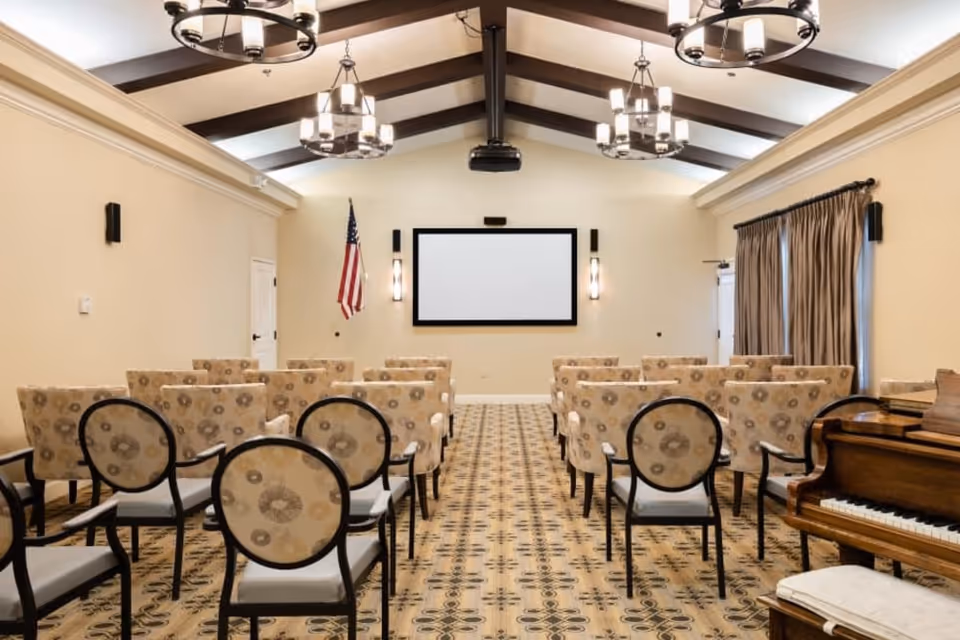 A bright meeting room with rows of upholstered chairs facing a wall-mounted projection screen, chandeliers overhead, an American flag, and a piano at the side.