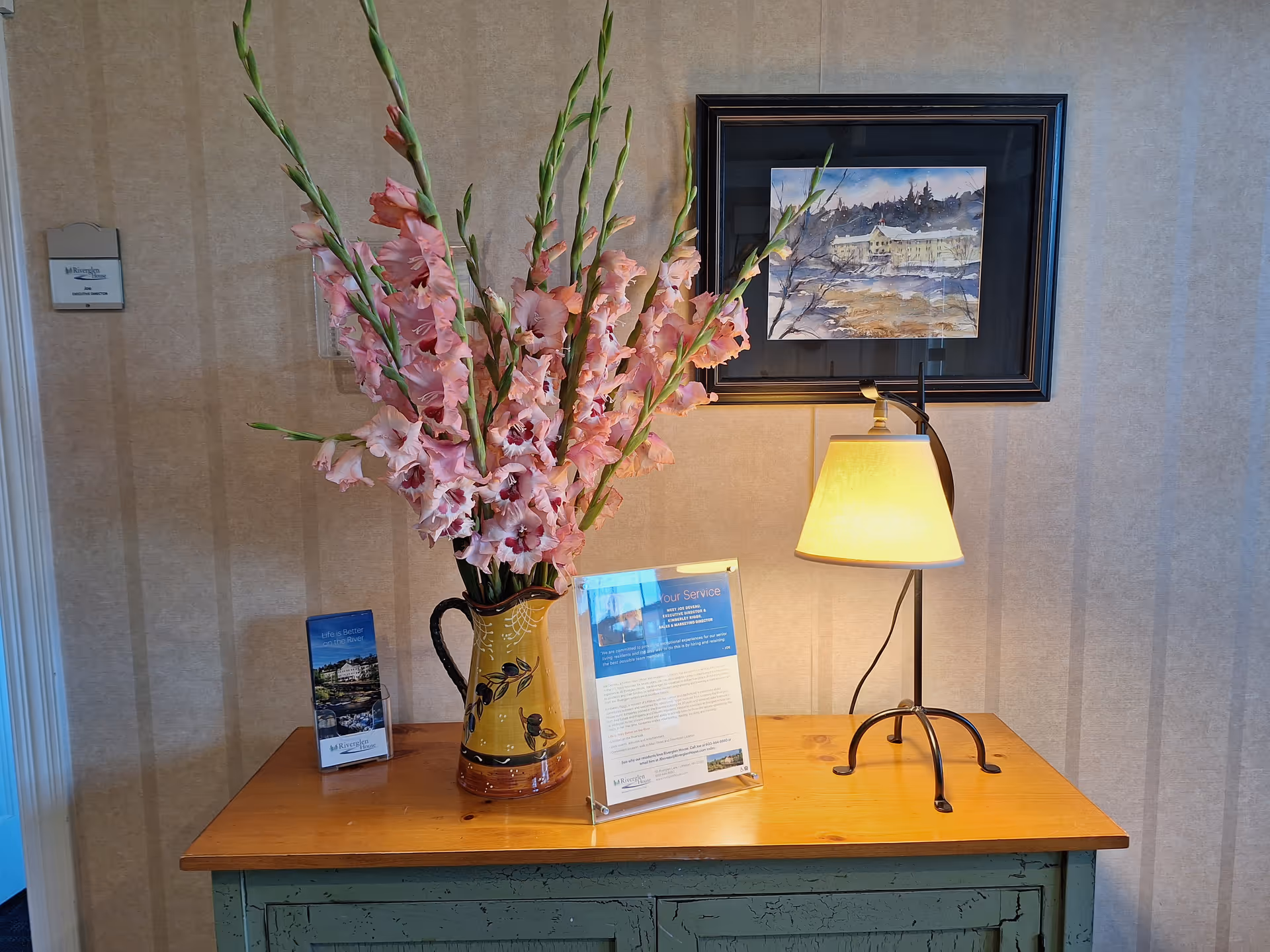 Wooden console table with a large vase of pink gladiolus, a lit lamp, framed artwork, and brochures against a wallpapered wall.