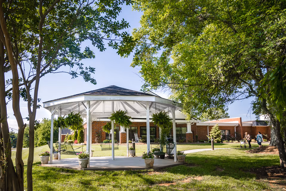 A white gazebo with hanging plants and swings in a grassy outdoor area surrounded by trees, with a brick building in the background and two people walking on a path nearby.