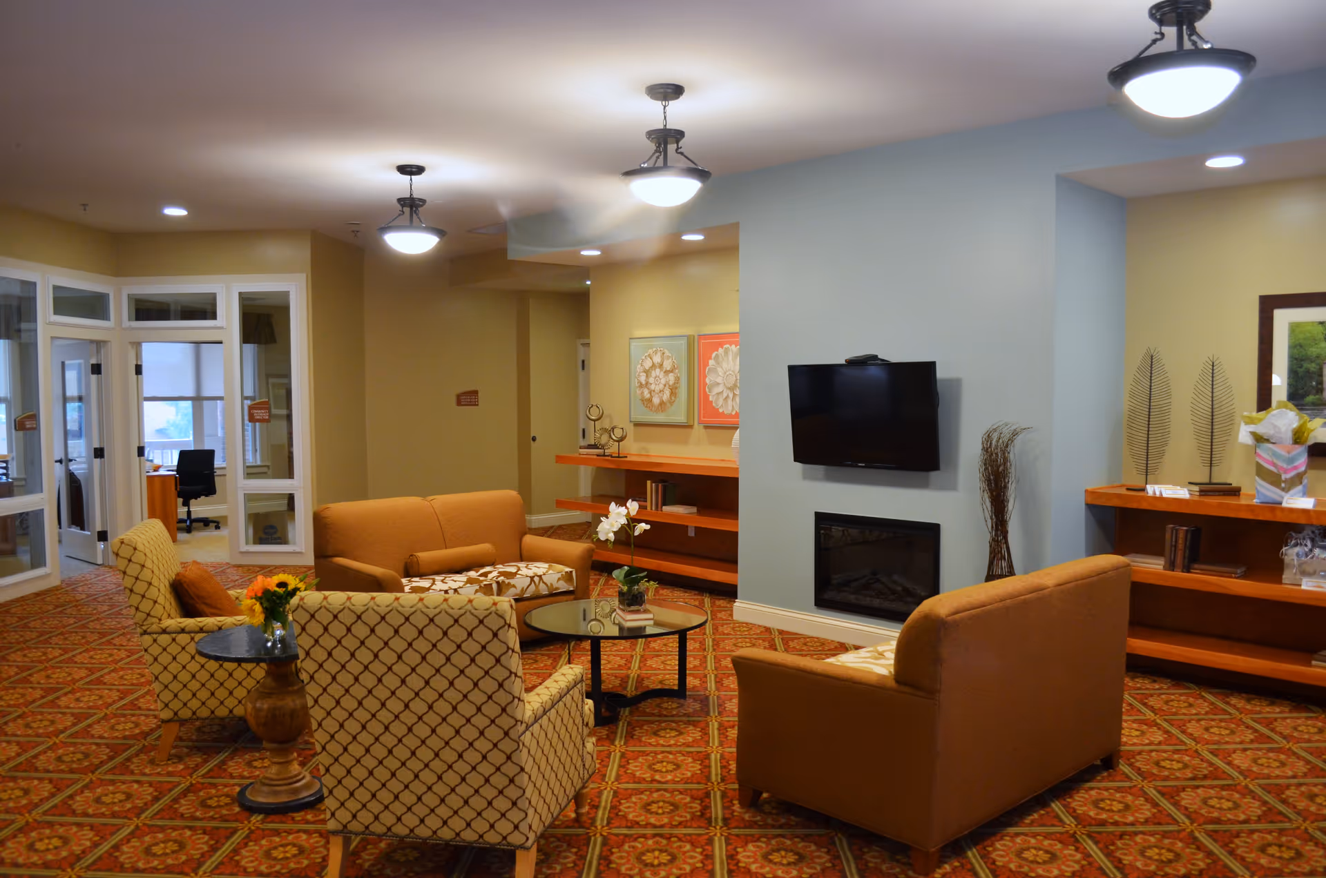 A cozy living room area in Pinnacle Place Memory Care featuring two patterned armchairs and two brown sofas arranged around a glass coffee table with a small plant and books. The room has a patterned carpet, a wall-mounted TV above a fireplace, and built-in wooden shelves with decorative items. The walls are painted in soft beige and light blue tones, and there are ceiling lights providing warm illumination.