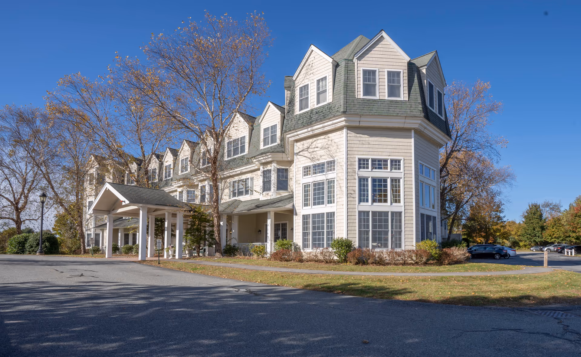 Exterior view of a multi-story assisted living facility building with large windows, a covered entrance, and surrounding trees under a clear blue sky.