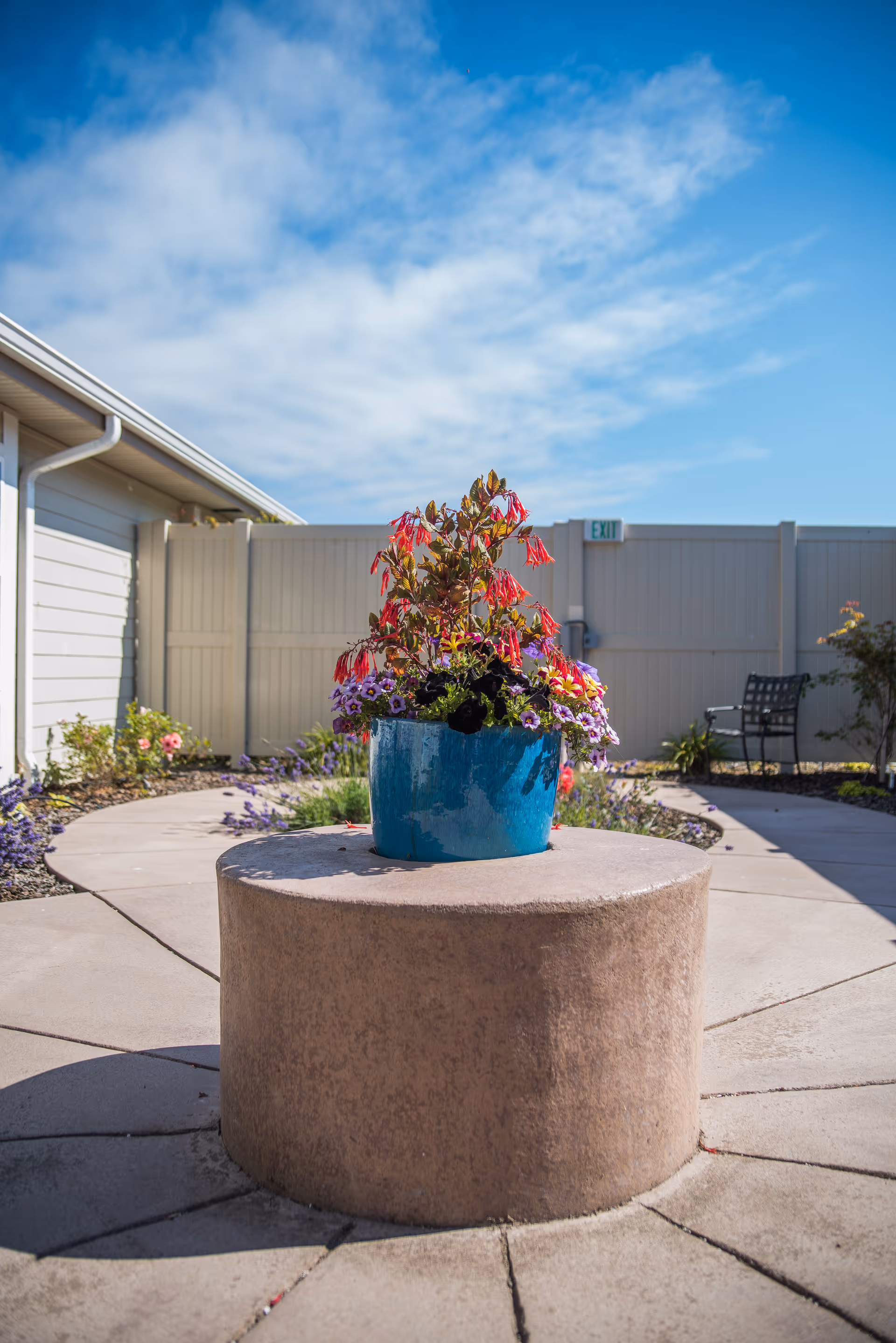 Outdoor patio area with a round concrete table in the center holding a blue pot filled with colorful flowers. The area is surrounded by a beige fence, some garden plants, and a metal chair. The sky is partly cloudy and blue.