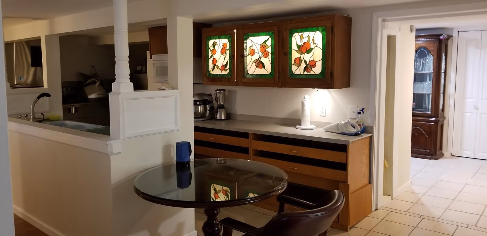 Interior view of a kitchen area with a round glass table and a chair in the foreground. The kitchen features wooden cabinets with stained glass panels, a countertop with kitchen appliances, and a pass-through window to another room. There is a hallway leading to another room with a wooden china cabinet and white double doors.