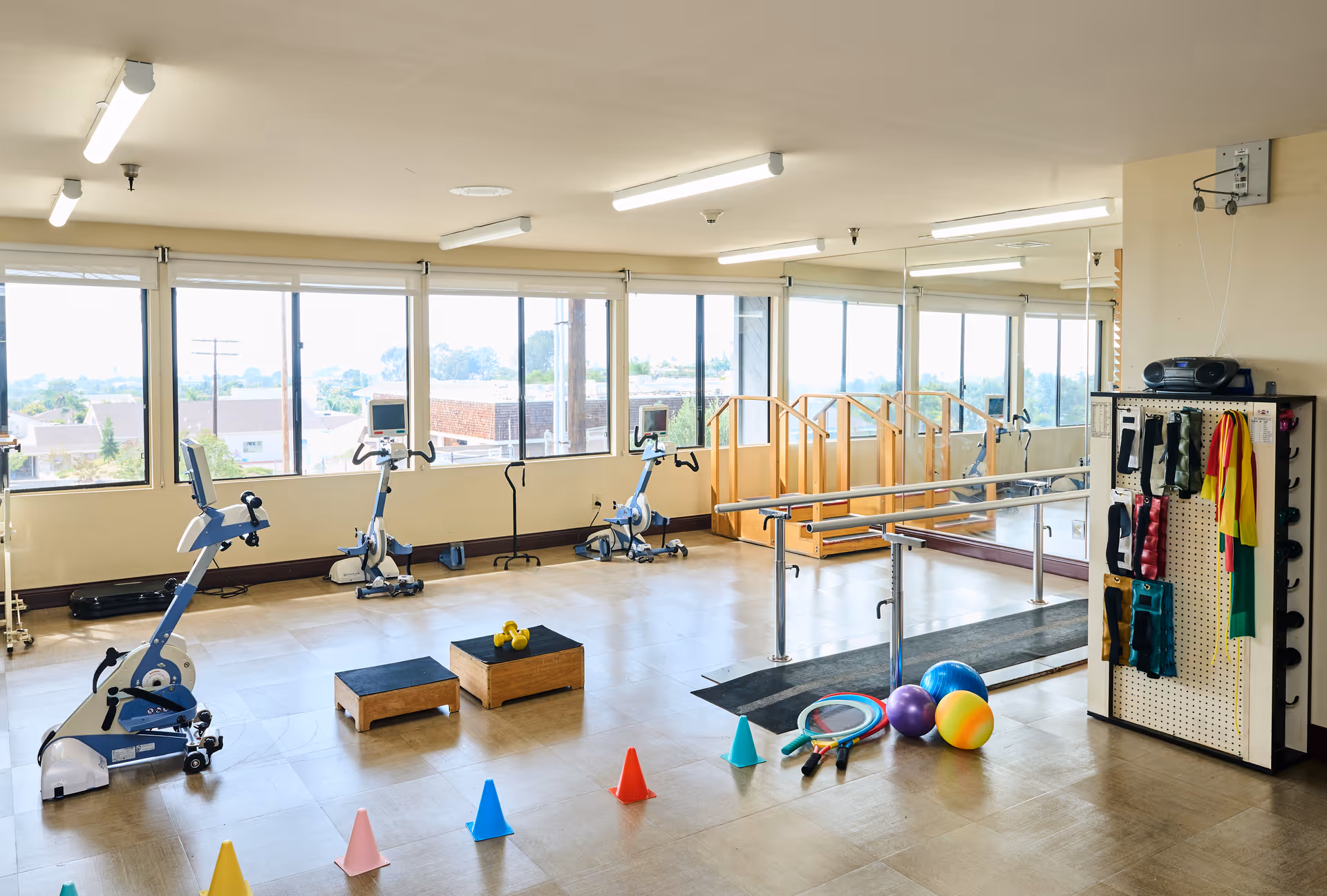 A bright rehabilitation exercise room with large windows, stationary exercise bikes, step platforms, colorful cones, exercise balls, resistance bands, and a parallel bar setup for physical therapy.