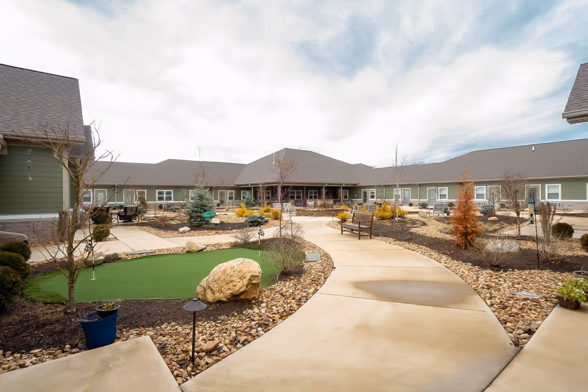 Outdoor courtyard area of Princeton Transitional Care featuring a winding concrete pathway, benches, landscaped garden beds with rocks and small trees, and a small artificial putting green. The courtyard is surrounded by single-story green buildings with gray roofs under a partly cloudy sky.