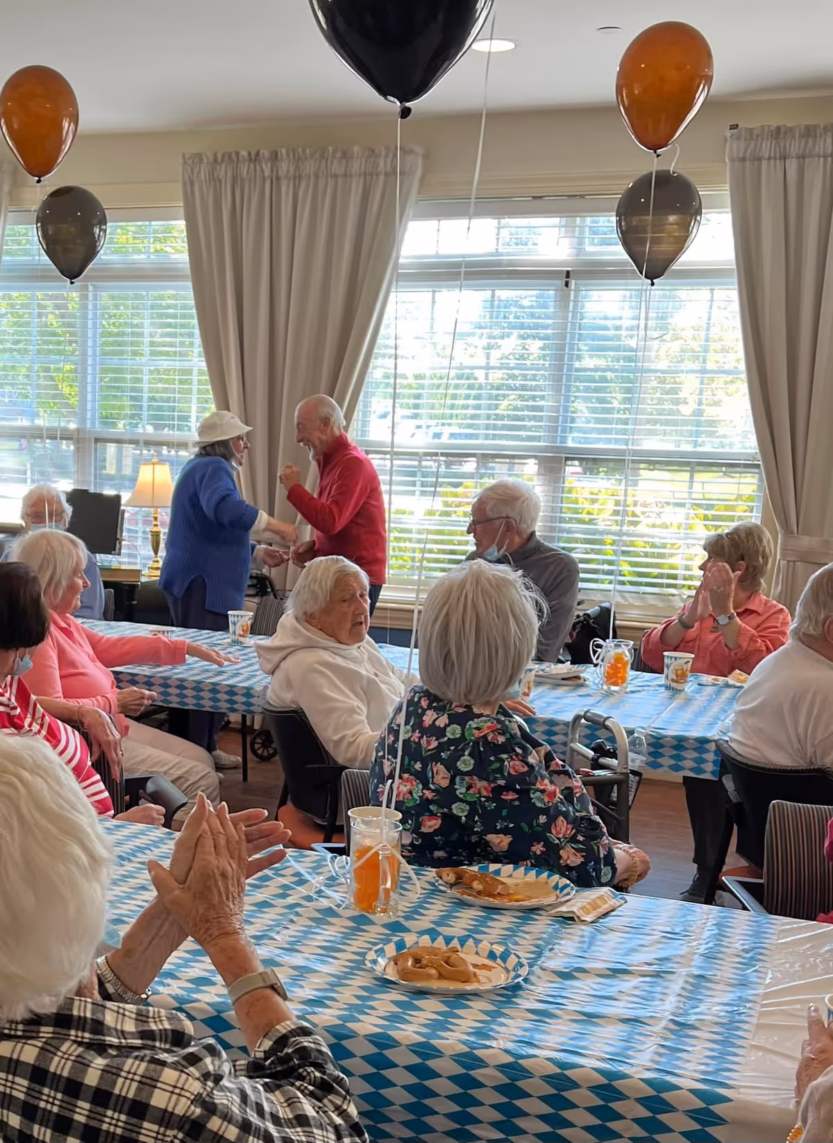 Elderly residents gathered around decorated tables in a sunlit dining/activity room with balloons.