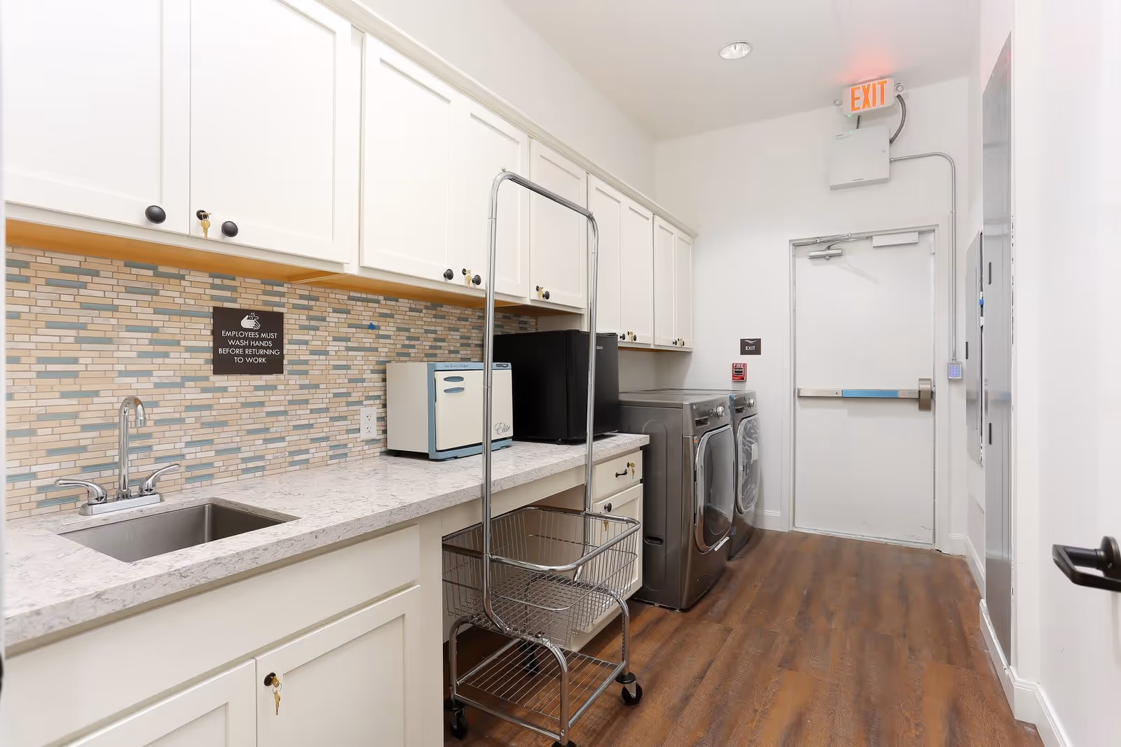 A clean laundry room with white cabinets, a countertop with a sink, a small appliance, a black mini fridge, a metal laundry cart, and a matching washer and dryer. The room has a tiled backsplash and wood flooring, with an exit door at the end.