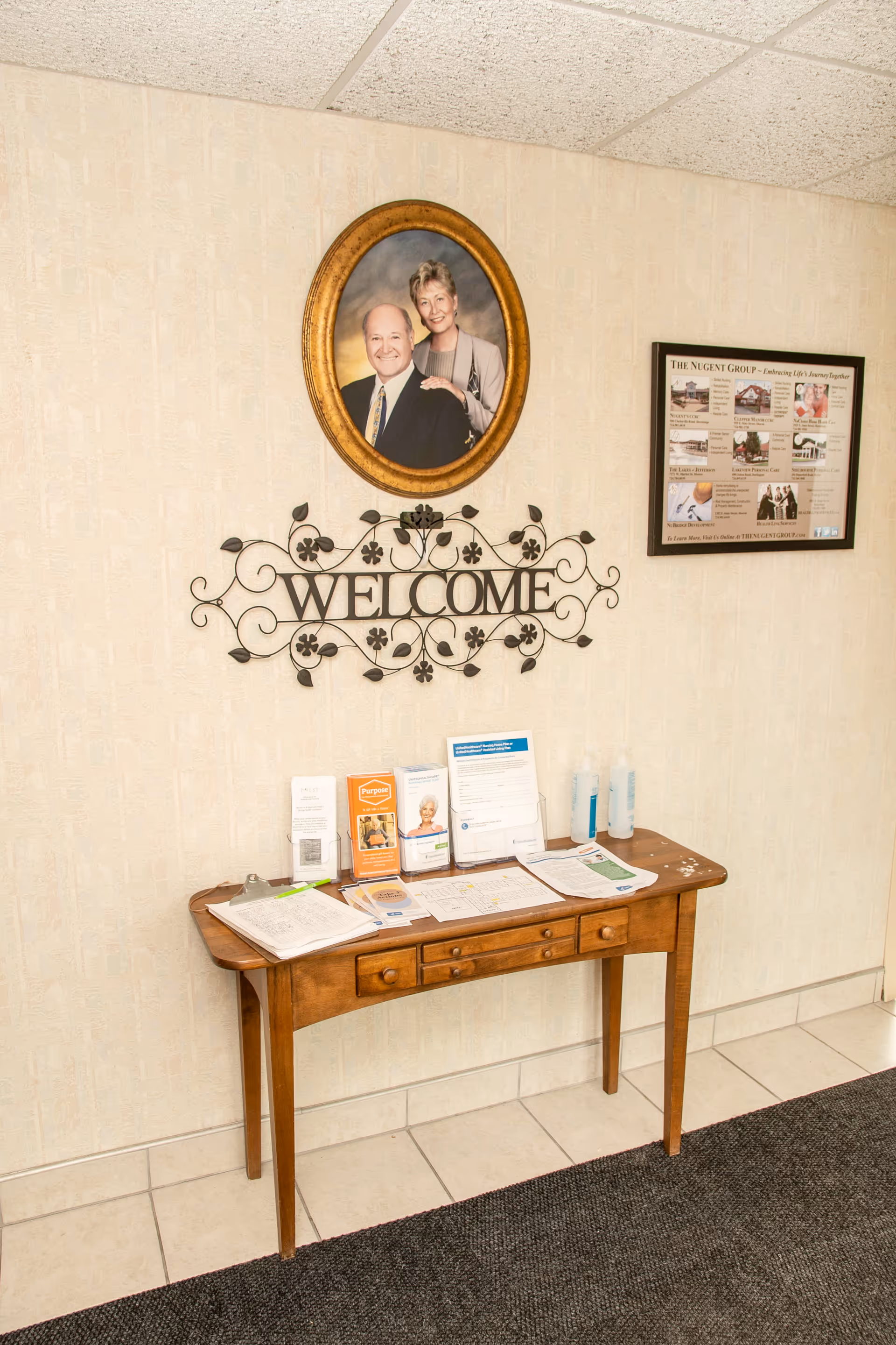 A wooden table against a beige wall with informational brochures, papers, and two bottles of hand sanitizer on top. Above the table is a decorative metal wall art piece with the word 'WELCOME' and a framed oval portrait of a smiling man and woman. To the right, there is a framed informational poster on the wall. The floor is tiled with a dark carpet in front of the table.