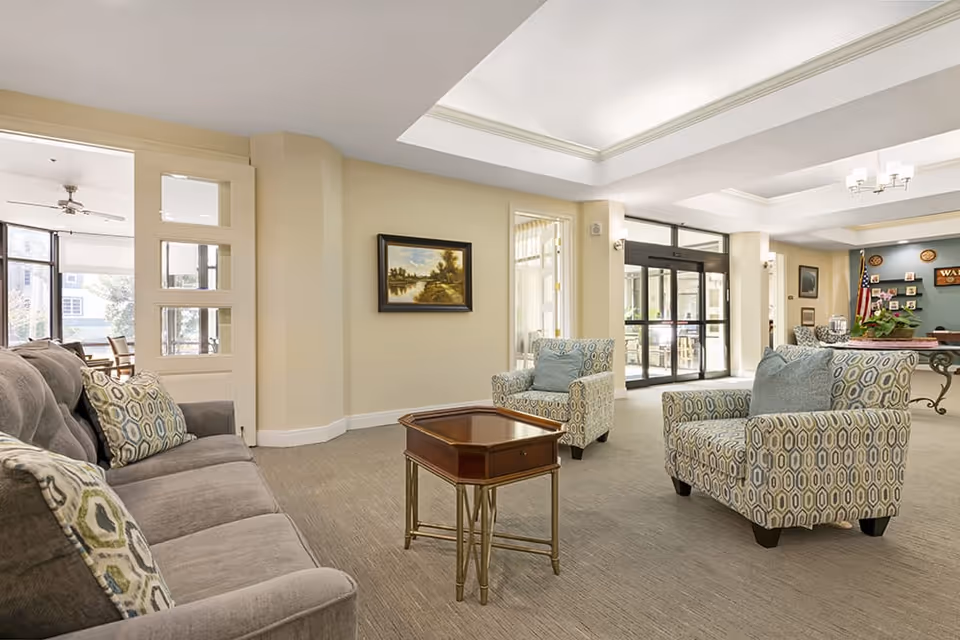 Bright furnished lounge area with sofas and patterned armchairs around a central wooden coffee table near large glass entrance doors.
