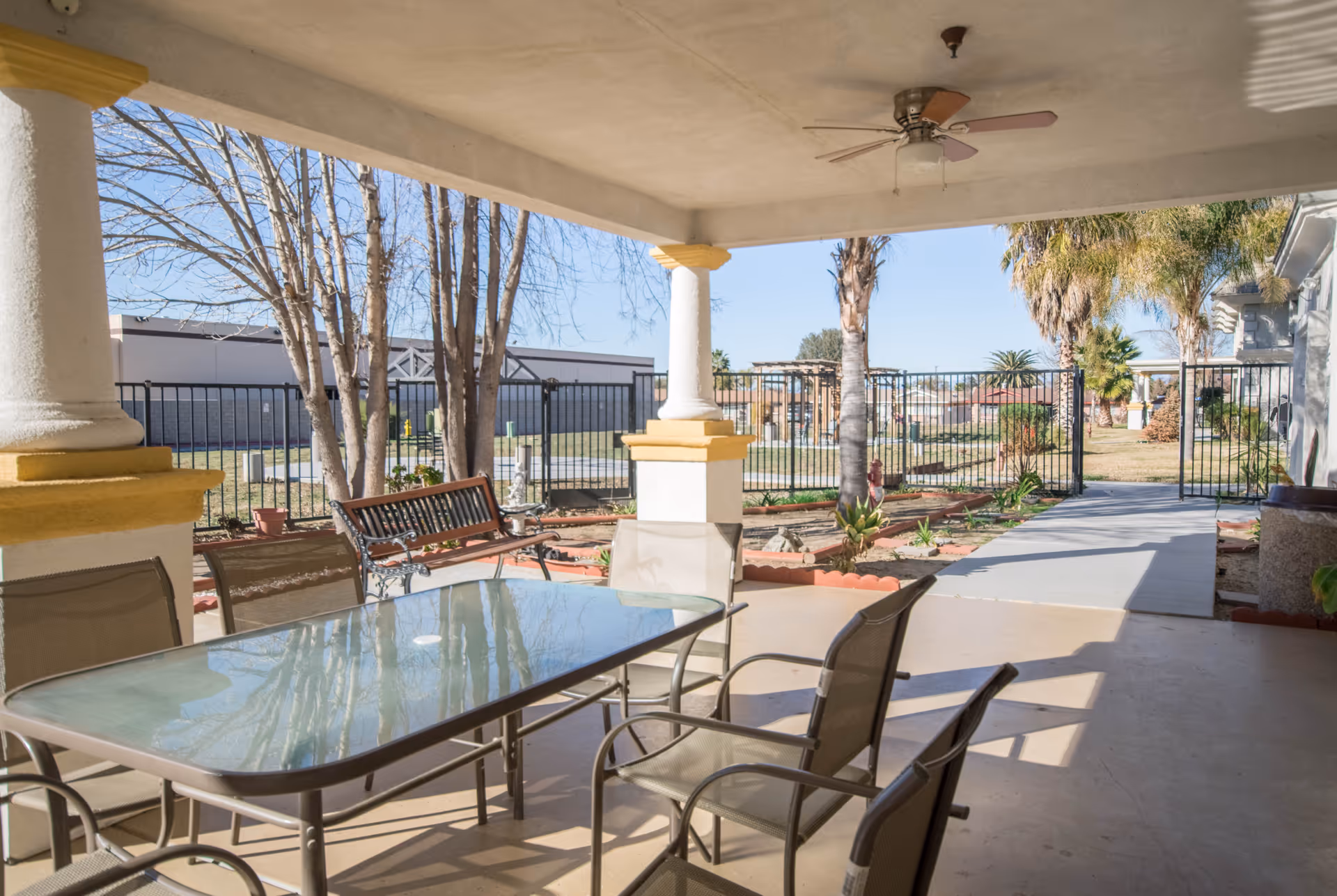 Covered patio with a glass-top table and chairs, ceiling fan, bench, and a view of a fenced yard and walkway.
