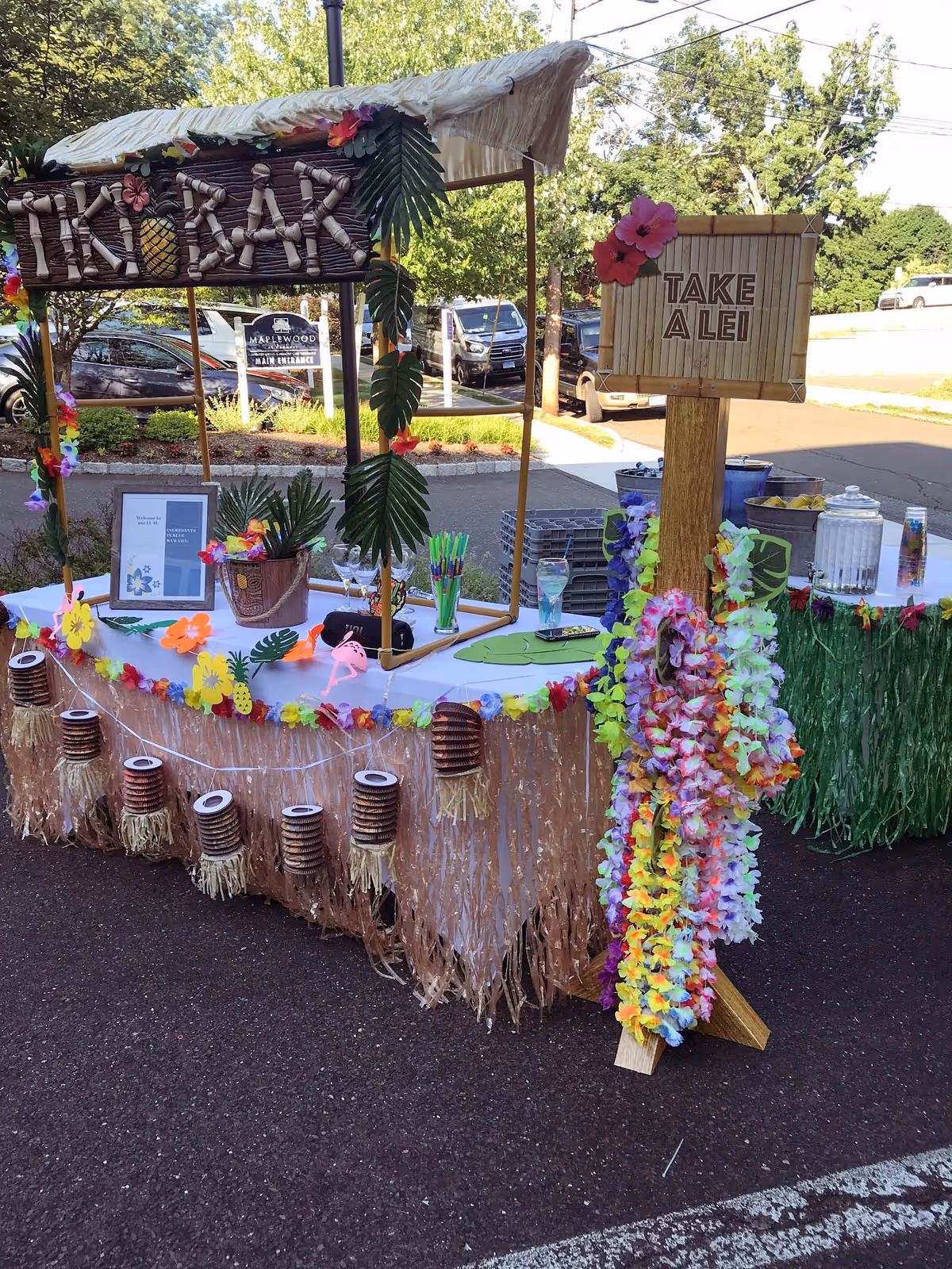 Outdoor tiki bar setup decorated with tropical flowers, grass skirts, and hanging lanterns. A wooden sign reads 'TAKE A LEI' with colorful leis hanging from it. In the background, there are parked cars and a sign for Maplewood at Danbury main entrance.