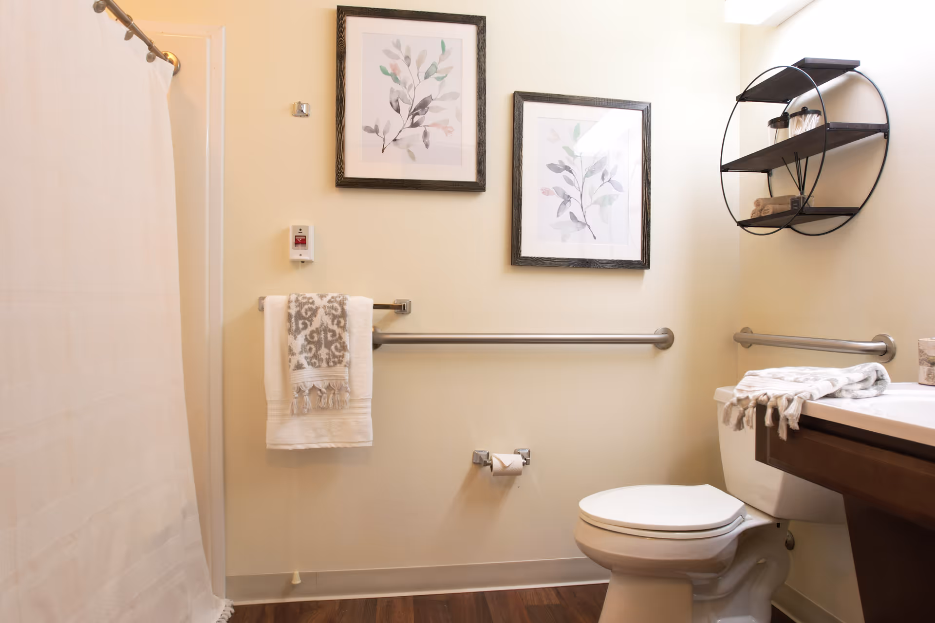 A clean and well-lit bathroom with a white toilet, a shower with a white curtain, a towel rack holding a white towel with a gray patterned towel on top, two framed botanical prints on the wall, a circular black metal shelf with towels and jars, and a wooden vanity with a white countertop.