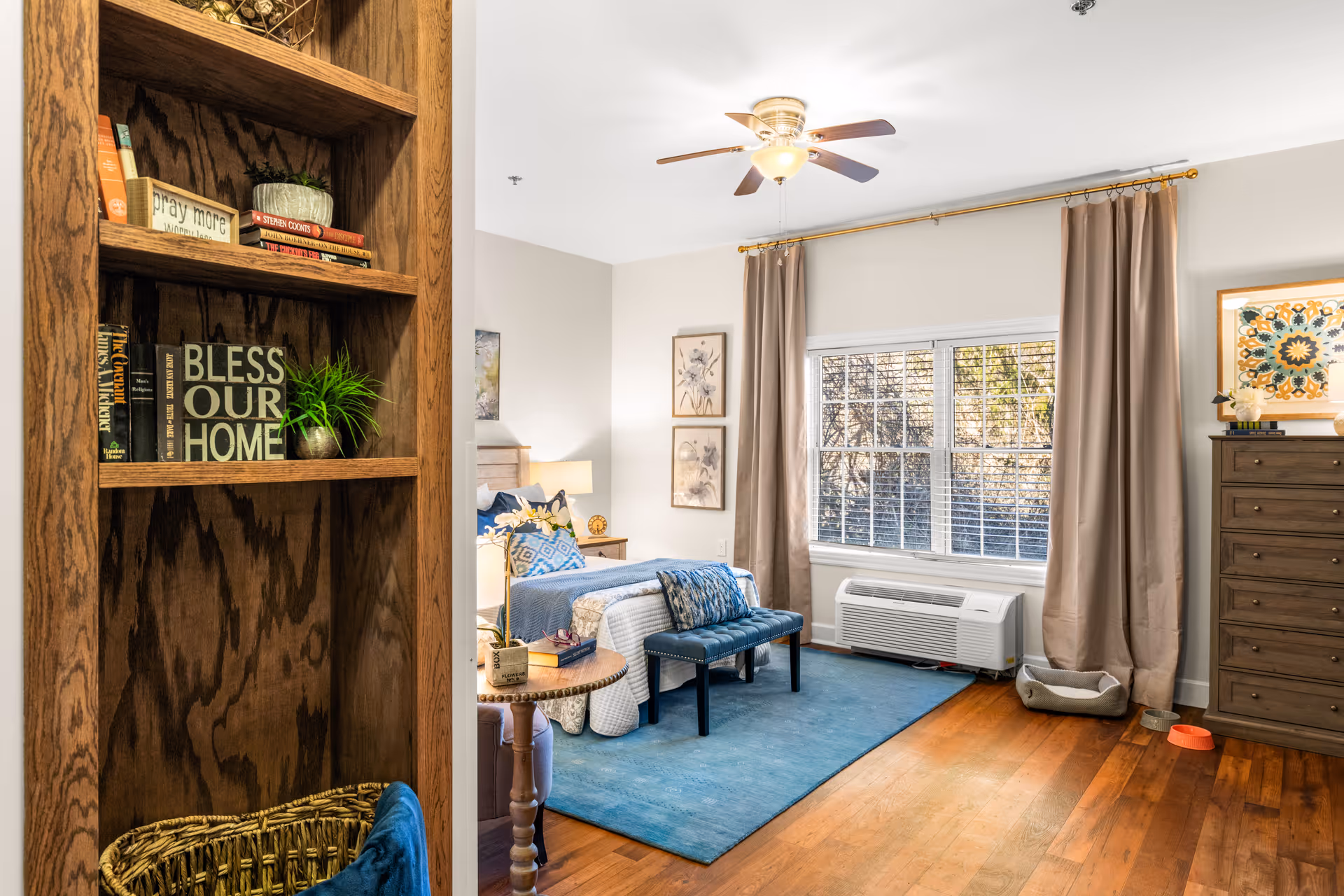 A bright and cozy bedroom in a senior living facility with a bed adorned with blue and white bedding, a blue bench at the foot of the bed, a wooden dresser, and a large window with beige curtains letting in natural light. A wooden bookshelf with books and decorative items is visible on the left side, and a ceiling fan with a light fixture is mounted on the white ceiling.