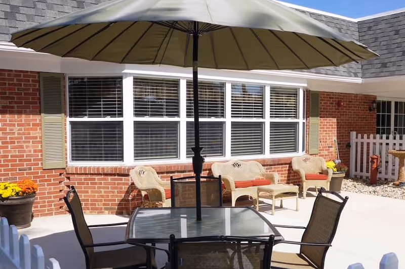 Outdoor patio area with a glass-top table and four chairs under a large umbrella. Behind the table, there are wicker chairs with red cushions and a small wicker table. The background features a brick building with white-framed windows and green shutters, along with potted flowers and a white picket fence.