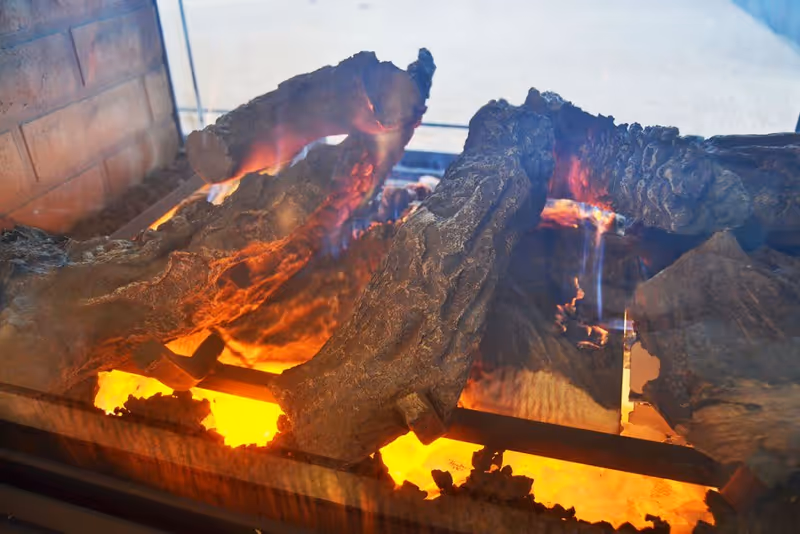 Close-up view of artificial logs burning in a gas fireplace with visible flames and glowing embers behind a glass screen.