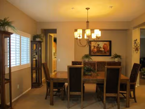 A warm dining room with a wooden table, chairs, chandelier, and decorative plants.