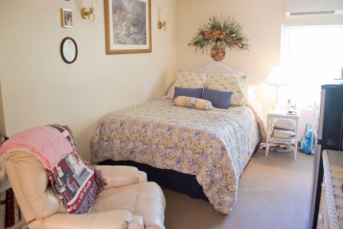 A cozy bedroom with a floral-patterned bed, an upholstered armchair, a nightstand with a lamp, and wall decorations.