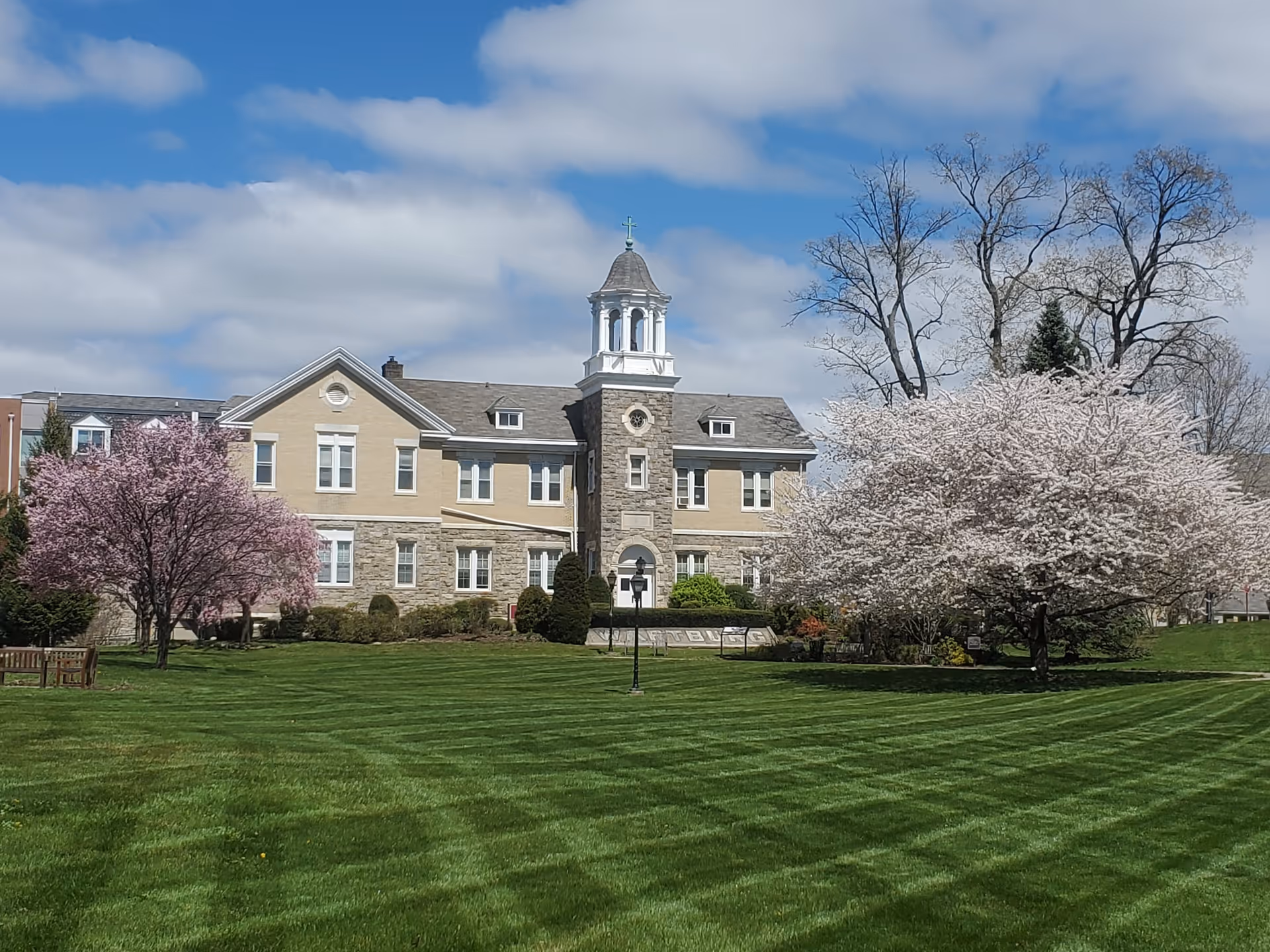 A large, stately building with a stone and beige facade, featuring a central tower with a cupola. The building is surrounded by a well-maintained lawn with striped mowing patterns, blossoming pink and white trees, and a clear blue sky with some clouds.