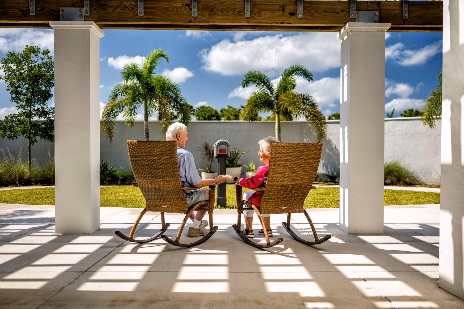 An elderly couple sitting on wicker rocking chairs under a shaded patio, holding hands and facing a garden with palm trees and a blue sky with clouds.