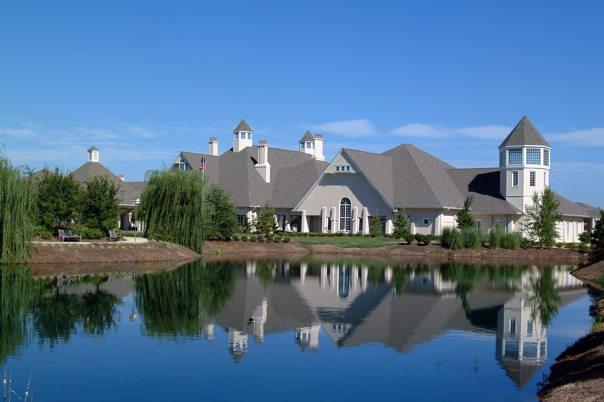 Exterior view of a large senior living facility building with multiple peaked roofs and a tower, reflected in a calm pond in front. The building is surrounded by trees and landscaping under a clear blue sky.