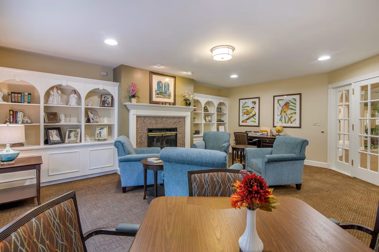 Bright senior living lounge with blue armchairs around a fireplace, built-in shelving, and a table with a vase of flowers.
