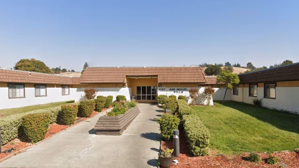 Single-story white brick building with a tiled roof and central glass entrance, a paved walkway and trimmed hedges in a courtyard under a clear sky.