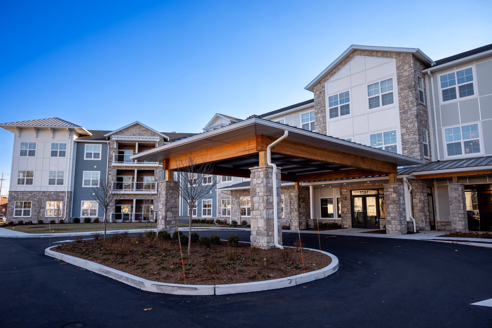 Exterior view of a multi-story senior living facility building with stone and siding facade, featuring a covered entrance with wooden beams and stone pillars, surrounded by a paved driveway and landscaped area with small trees and shrubs under a clear blue sky.