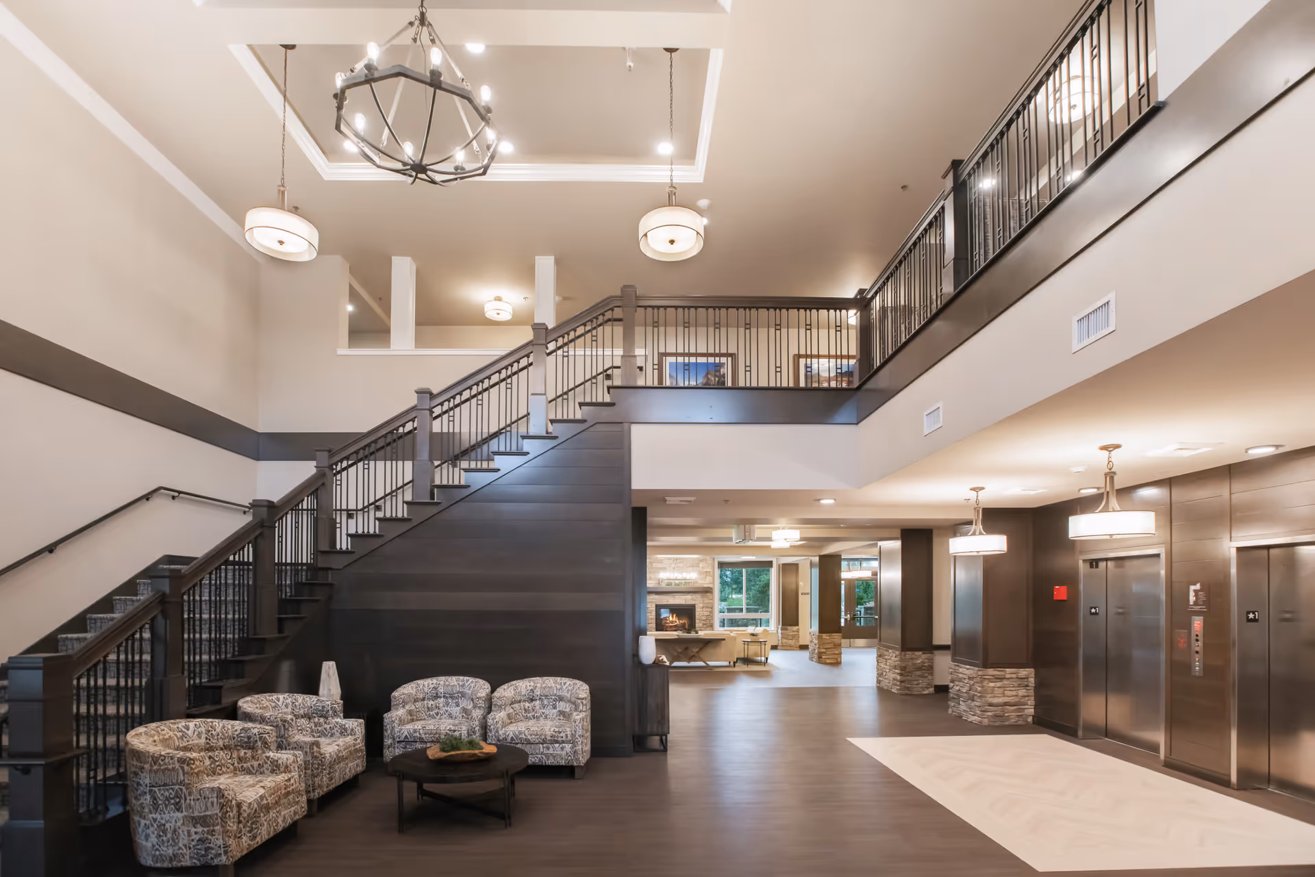 Spacious and modern interior of a senior living facility featuring a seating area with four patterned armchairs around a round coffee table, a staircase with dark wood and metal railings leading to an upper floor, and elevators on the right side. The space is well-lit with ceiling lights and has a neutral color palette with beige walls and dark wood accents.