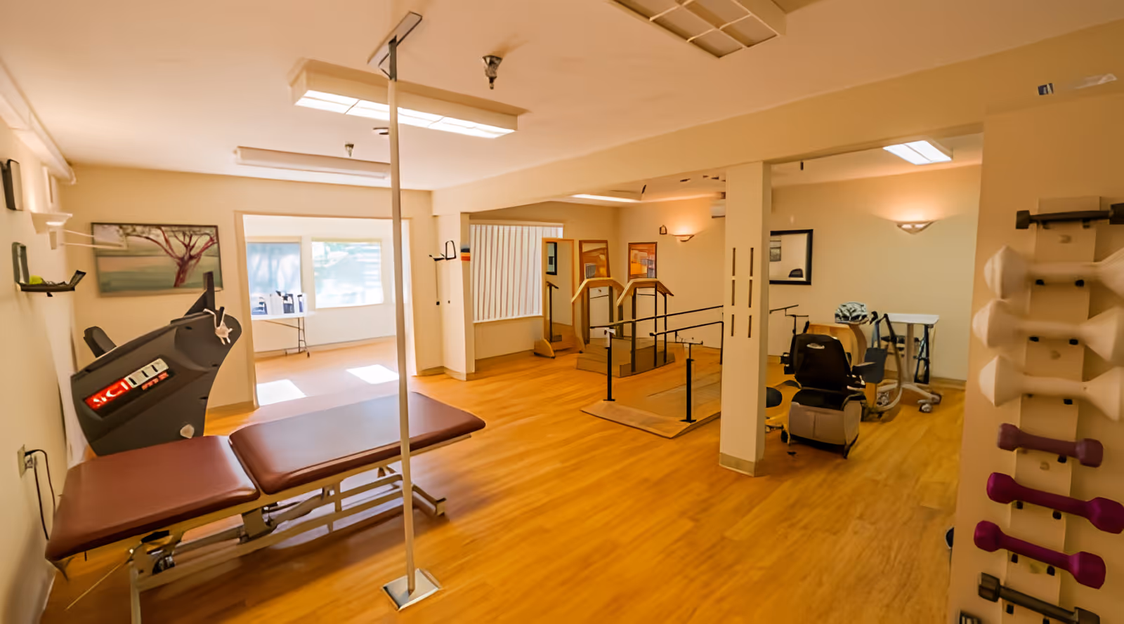 A rehabilitation therapy room with wooden flooring, featuring a padded treatment table, parallel bars for walking exercises, a stationary exercise bike, and a rack with various hand weights. The room is well-lit with ceiling lights and has a large window letting in natural light.