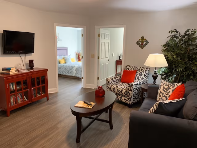 A cozy living room with a dark gray sofa and a patterned armchair, both adorned with red and black cushions. A wooden coffee table with a book and decorative item sits in the center. A red wooden cabinet with books and a TV mounted above it is on the left. Two doorways lead to a bedroom with a bed and a small hallway. A floor lamp and a large green plant add warmth to the space.