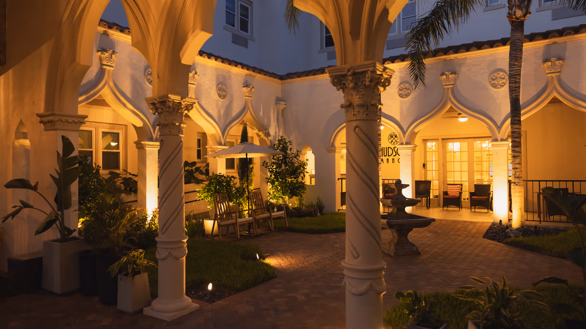 Evening view of an elegant courtyard at Hudson Manor Assisted Living, featuring arched columns, warm lighting, potted plants, a central fountain, and seating areas with chairs and umbrellas.