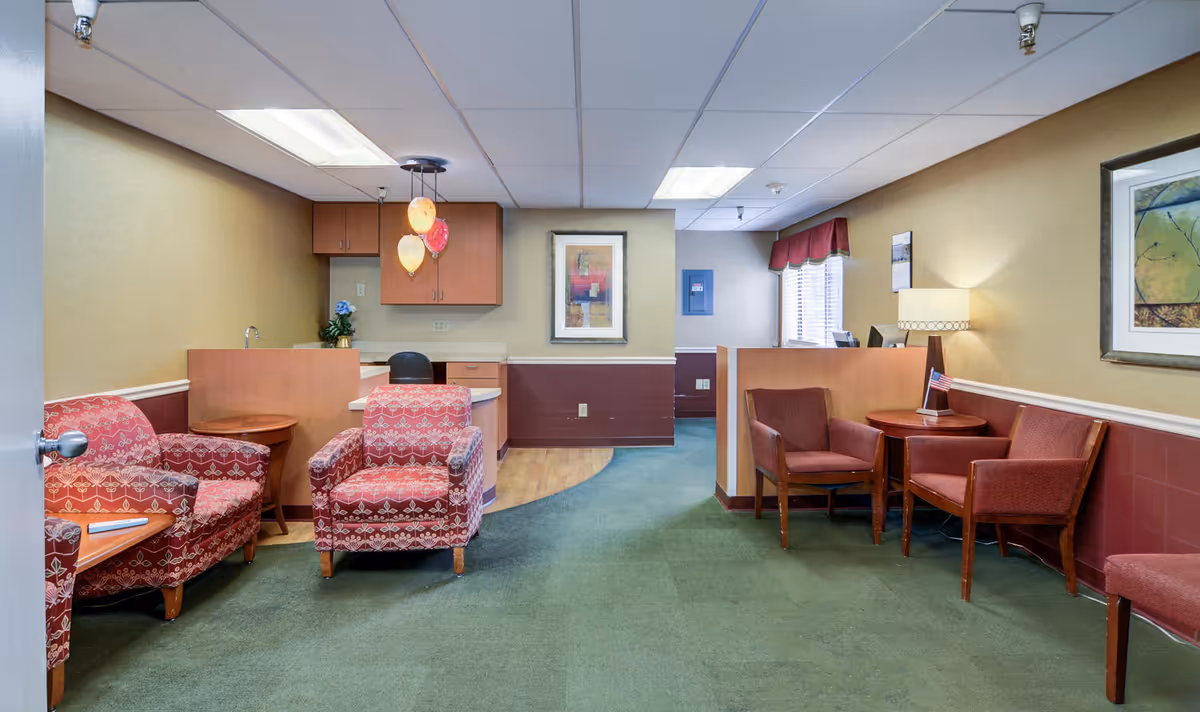 A senior living reception/waiting area with patterned armchairs, small tables, a reception desk and framed artwork on the walls.