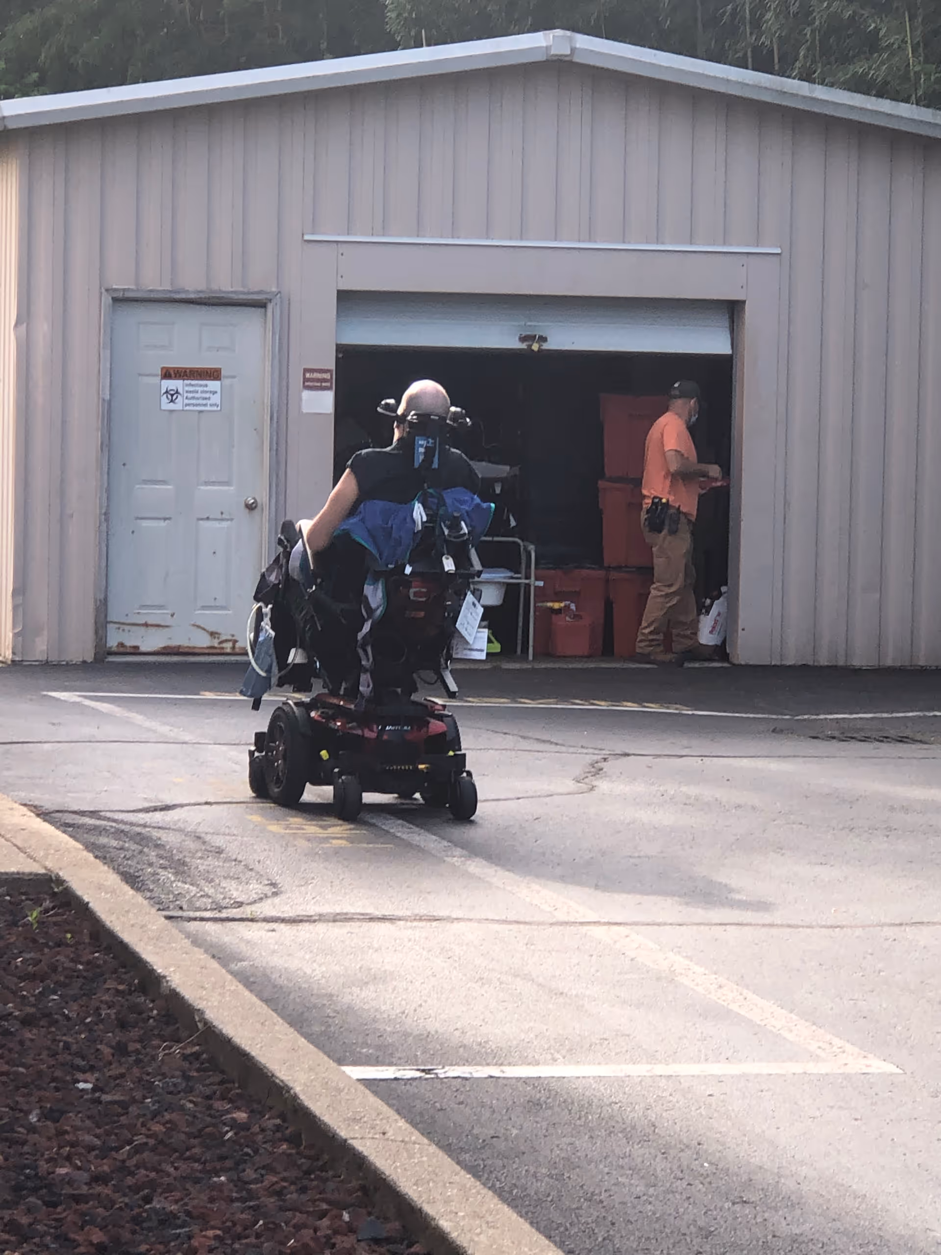 A person in a motorized wheelchair is moving towards a metal storage building with an open garage door. Another person is standing inside the building near red storage bins. The building has a closed white door with warning signs on it.
