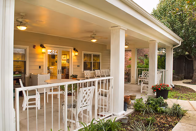Covered outdoor patio area with white railings and several white chairs and tables. The patio is attached to a building with double glass doors and windows, illuminated by wall-mounted lantern-style lights. There are plants and greenery along the edge of the patio and a tree in the background.