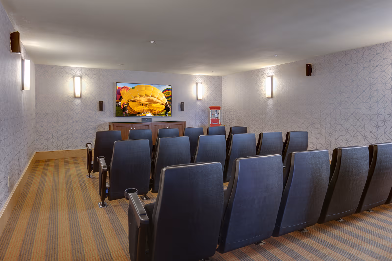A small theater room with three rows of black cushioned chairs facing a flat-screen TV mounted on a wall. The TV displays an image of a yellow hot air balloon being inflated. The room has patterned wallpaper, wall-mounted lights, and a popcorn machine on a cabinet beneath the TV.