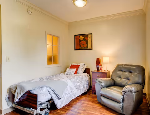 A cozy room with a single bed covered in a white and gray floral bedspread, a gray blanket folded at the foot of the bed, a wooden nightstand with a lamp and decorative items, a comfortable gray recliner chair, and a framed floral painting on the wall.