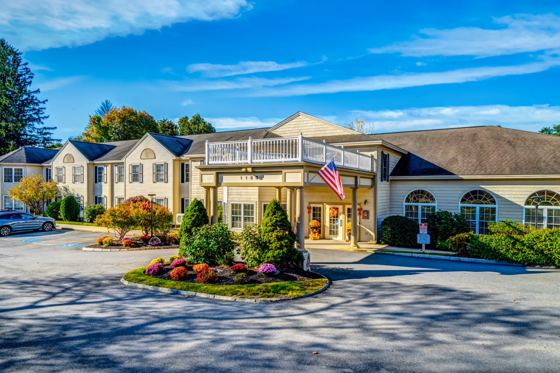 Front exterior of a senior living building with a covered entrance, American flag, and landscaped circular driveway.