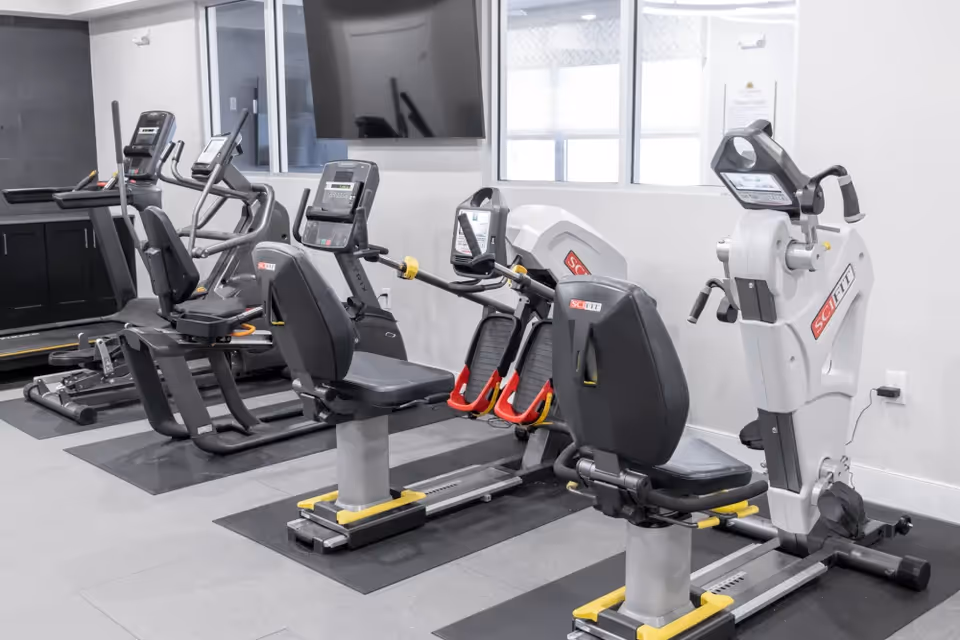 Interior view of a fitness room with exercise equipment including recumbent bikes, a rowing machine, and treadmills. The room has a large window, a wall-mounted TV, and light-colored walls.