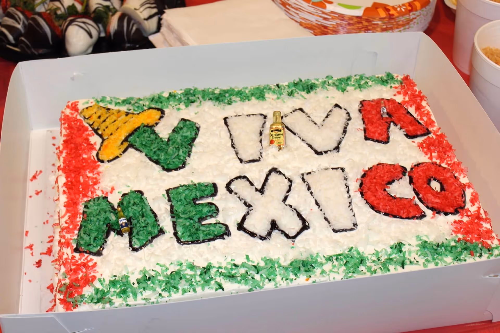 A rectangular cake decorated with colored coconut flakes in green, white, and red to resemble the Mexican flag. The cake has the words 'VIVA MEXICO' written on it with some letters filled in green and red, and a yellow sombrero decoration on the top left. There are two miniature bottles of alcohol placed on the cake. The cake is inside a white box with some chocolate-covered strawberries and white cups visible in the background.
