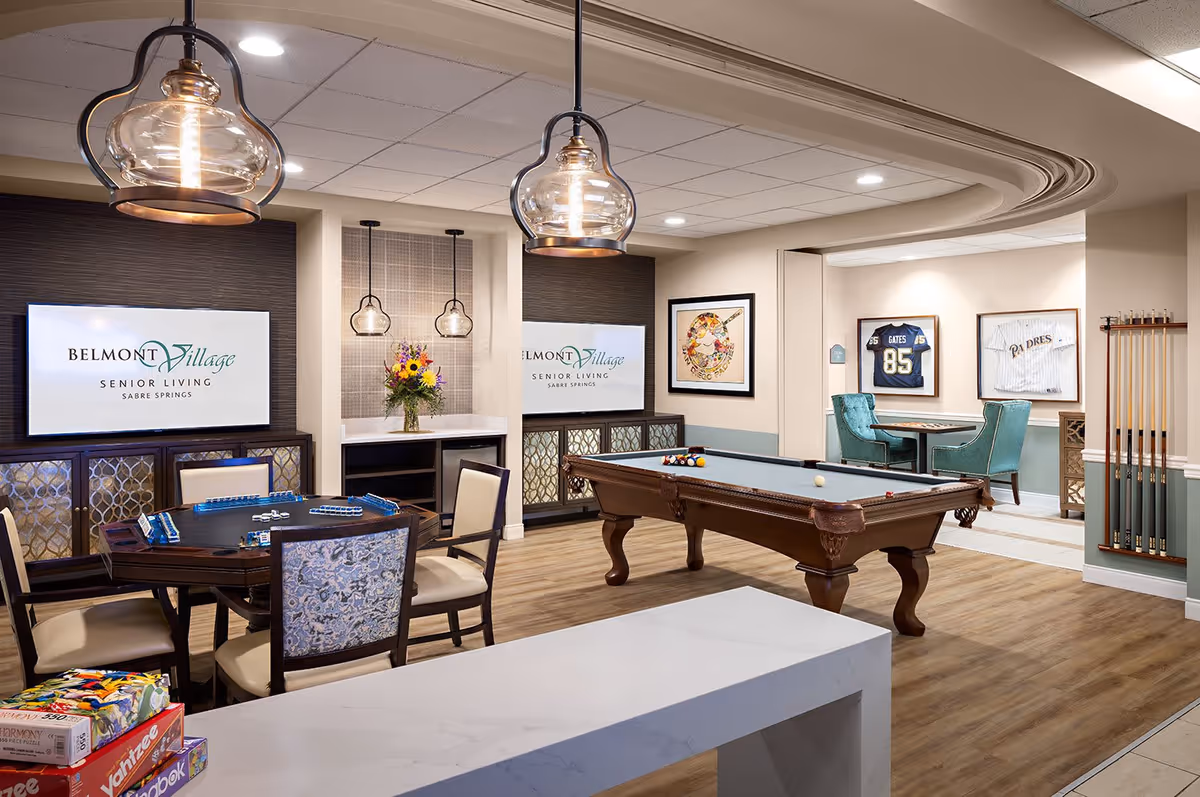 A recreational room in Belmont Village Senior Living Sabre Springs featuring a pool table with balls arranged for a game, a card table with chairs, and a small seating area with two teal chairs and a table. The room has wood flooring, pendant lights, framed sports jerseys on the wall, and a bouquet of flowers on a counter beneath two hanging lights. Two screens display the Belmont Village Senior Living Sabre Springs logo.