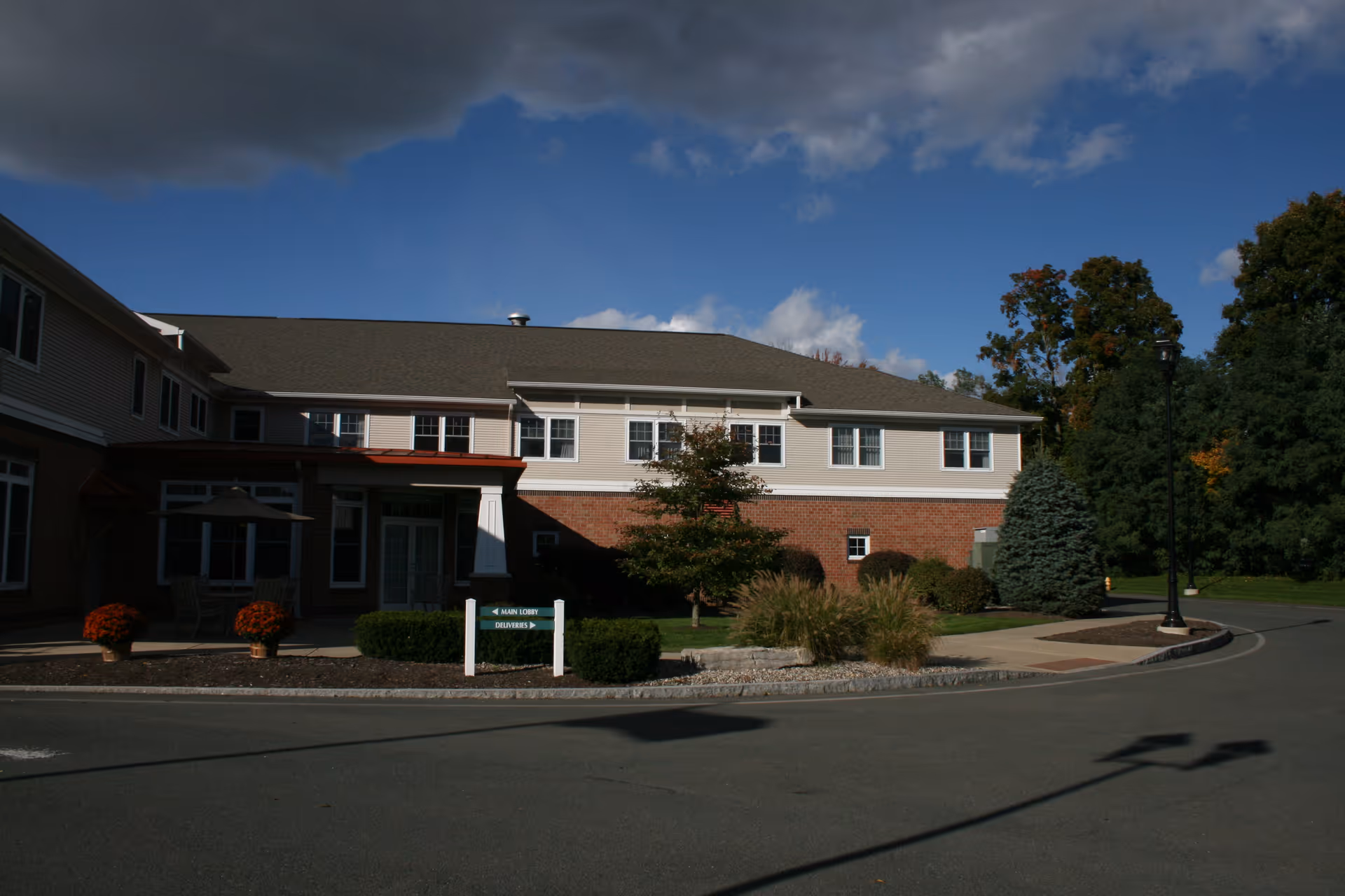 Front exterior of a two-story senior living building with a driveway, landscaping, and an entrance sign under a partly cloudy sky.