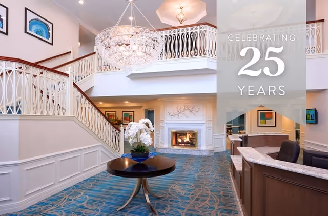 Interior view of a senior living facility lobby with a large chandelier, a staircase with white railings, a round table with a flower arrangement, a fireplace, and a reception desk. The area has blue patterned carpet and framed artwork on the walls. Text overlay reads 'Celebrating 25 Years'.