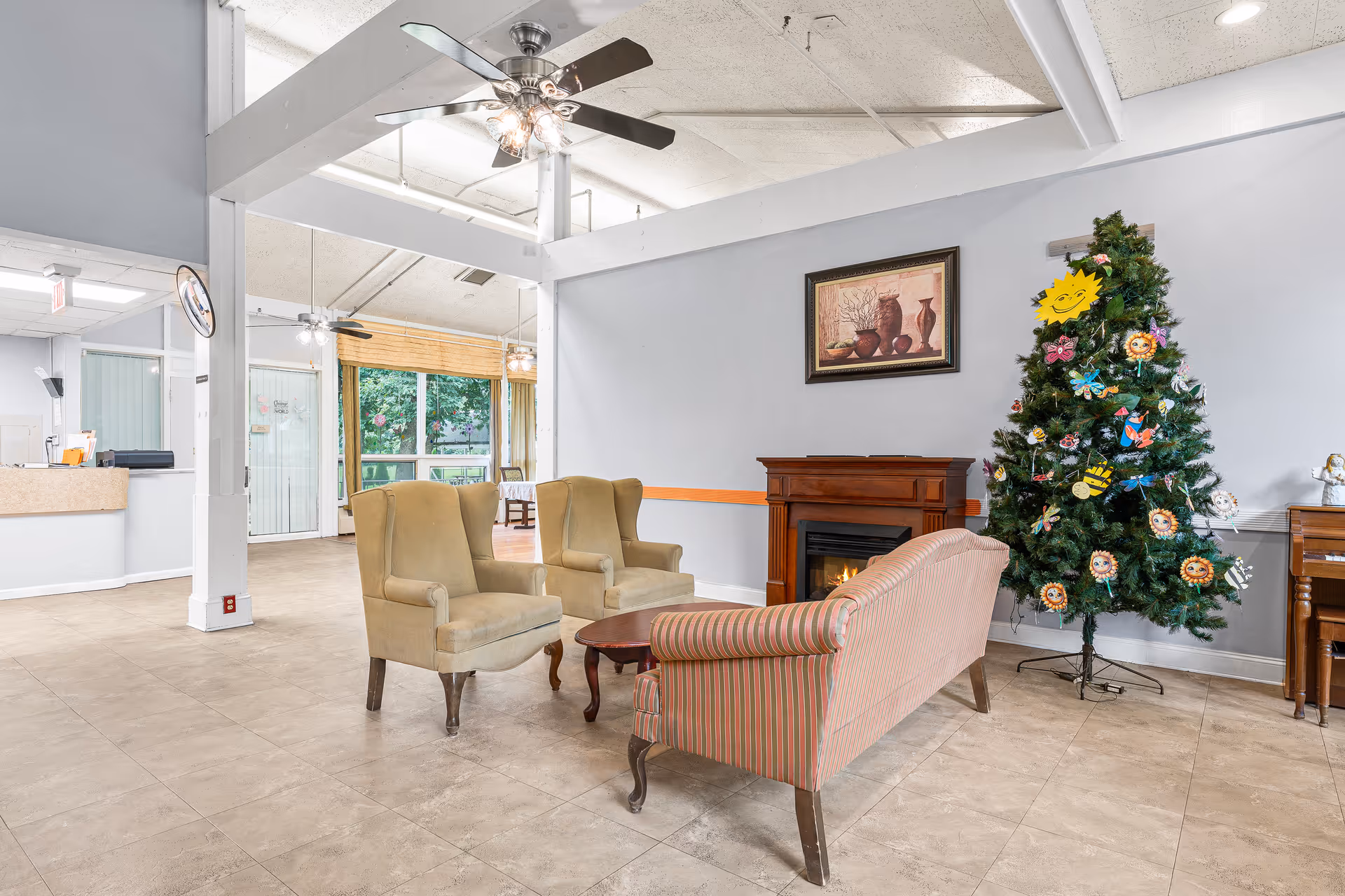 A cozy common area in a senior living facility featuring two beige armchairs and a striped sofa arranged around a small wooden coffee table. There is a decorative fireplace with a framed painting above it on the wall. To the right, a Christmas tree decorated with colorful handmade ornaments stands next to a piano. The room has tiled flooring, light gray walls, and a ceiling fan with lights. Large windows in the background let in natural light.
