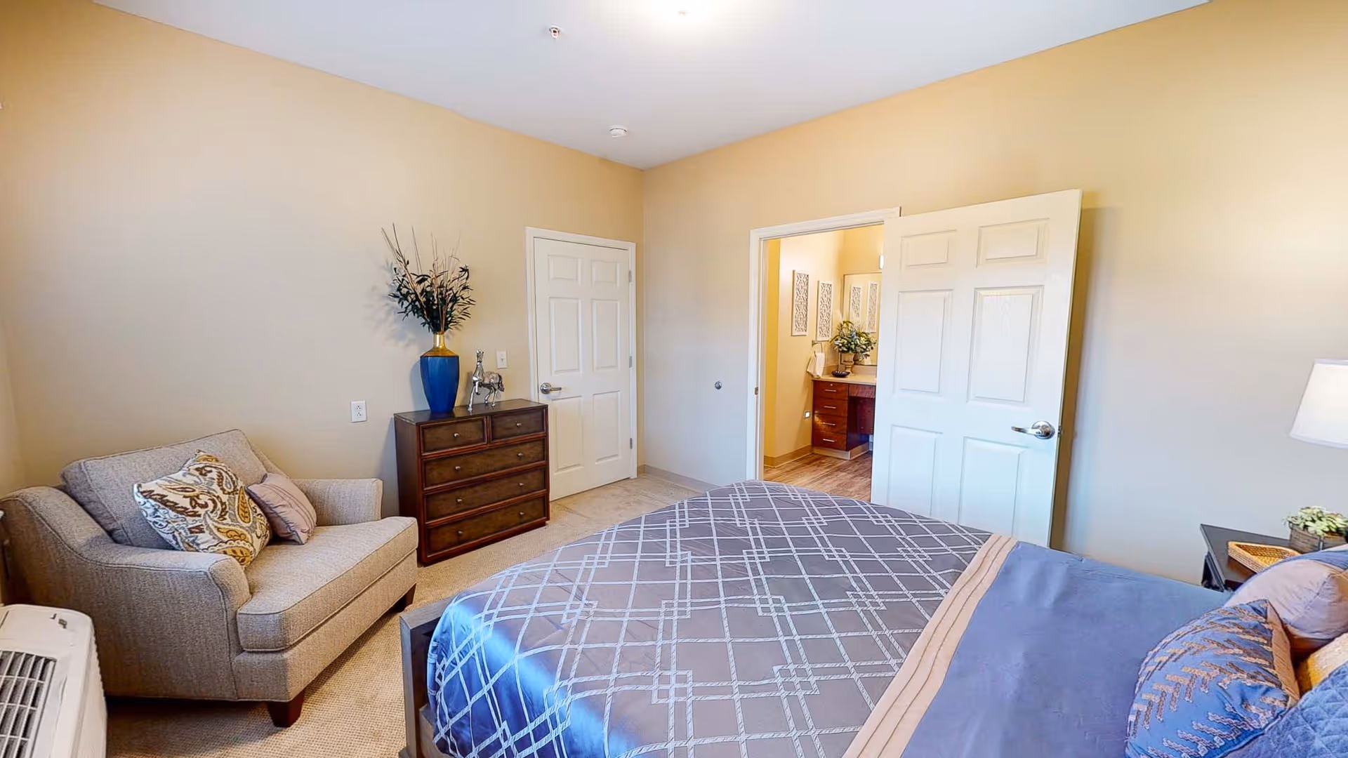 A bedroom with a bed covered in a blue and gray patterned comforter, a beige armchair with decorative pillows, a wooden dresser with a blue vase and decorative items, and an open door leading to a bathroom with a vanity and mirror.