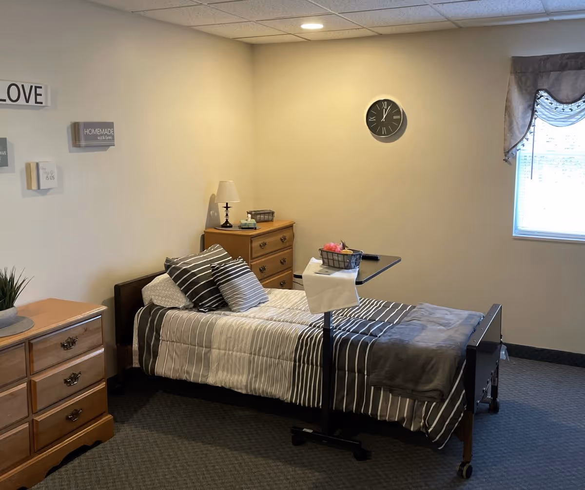 A neatly arranged bedroom in an assisted living facility featuring a single bed with striped bedding and multiple pillows. There is a wooden dresser with a lamp and tissue box on top, a small rolling overbed table with a basket, and a window with a valance letting in natural light. The walls are decorated with small framed signs and a clock.